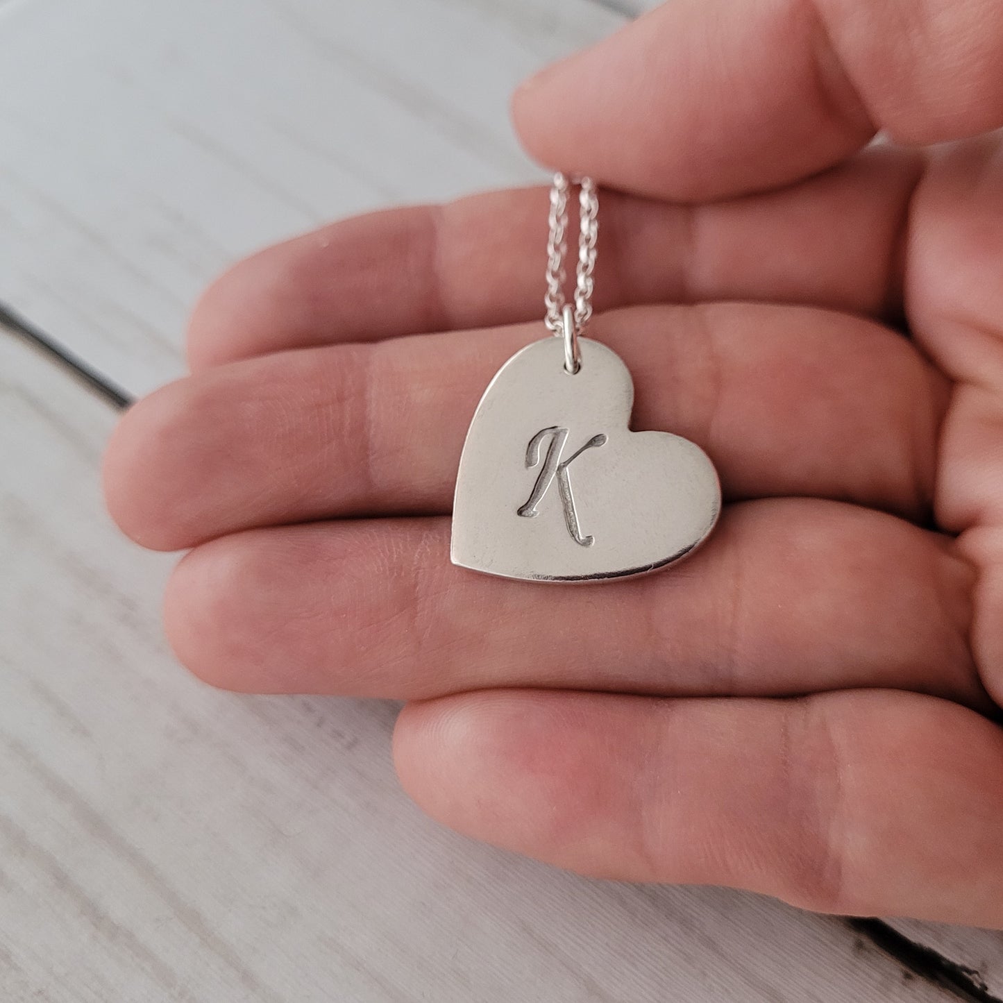 Silver heart-shaped pendant with 'K' held in a hand on a light wooden surface