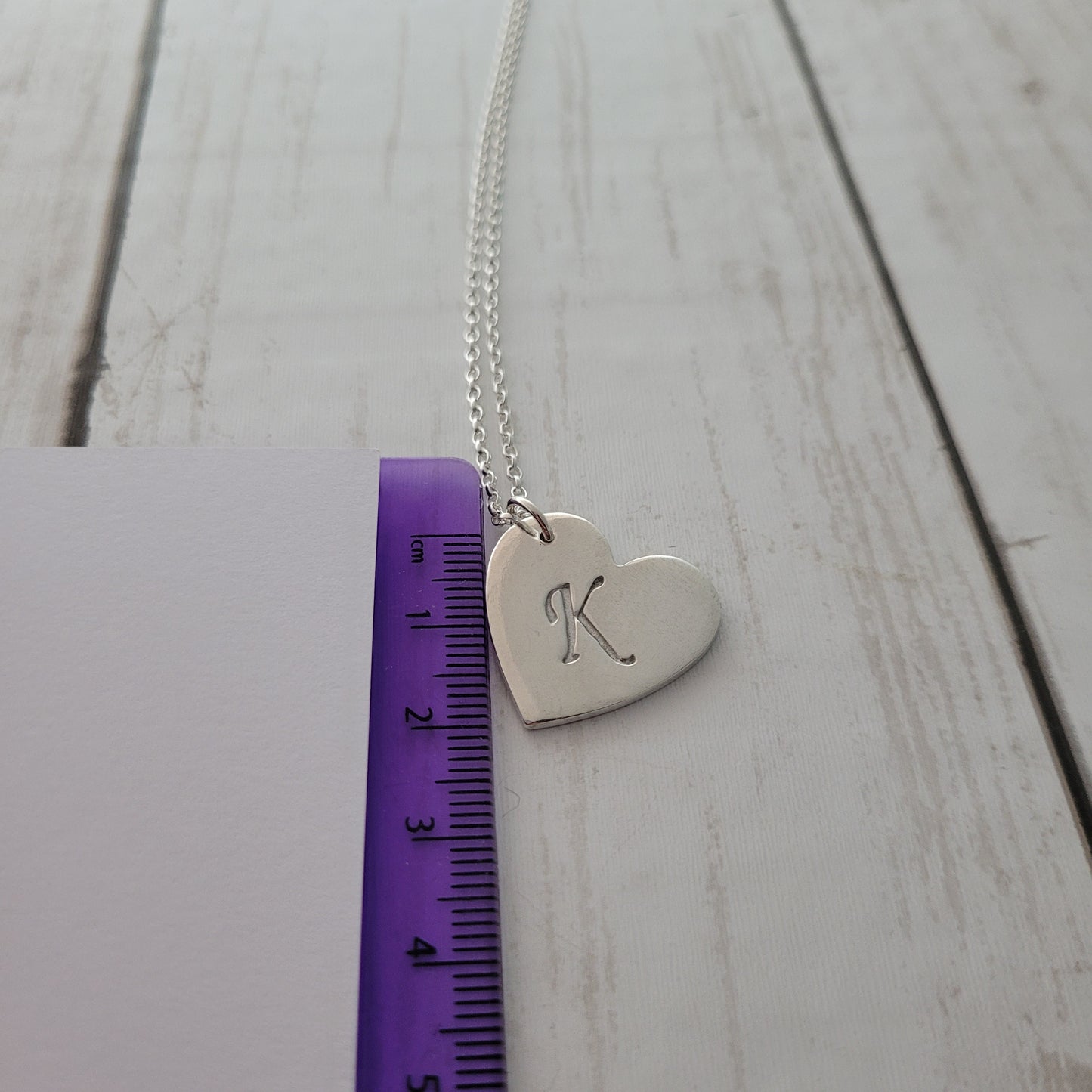 Silver heart-shaped pendant with engraved design on a wooden surface with a ruler for scale.