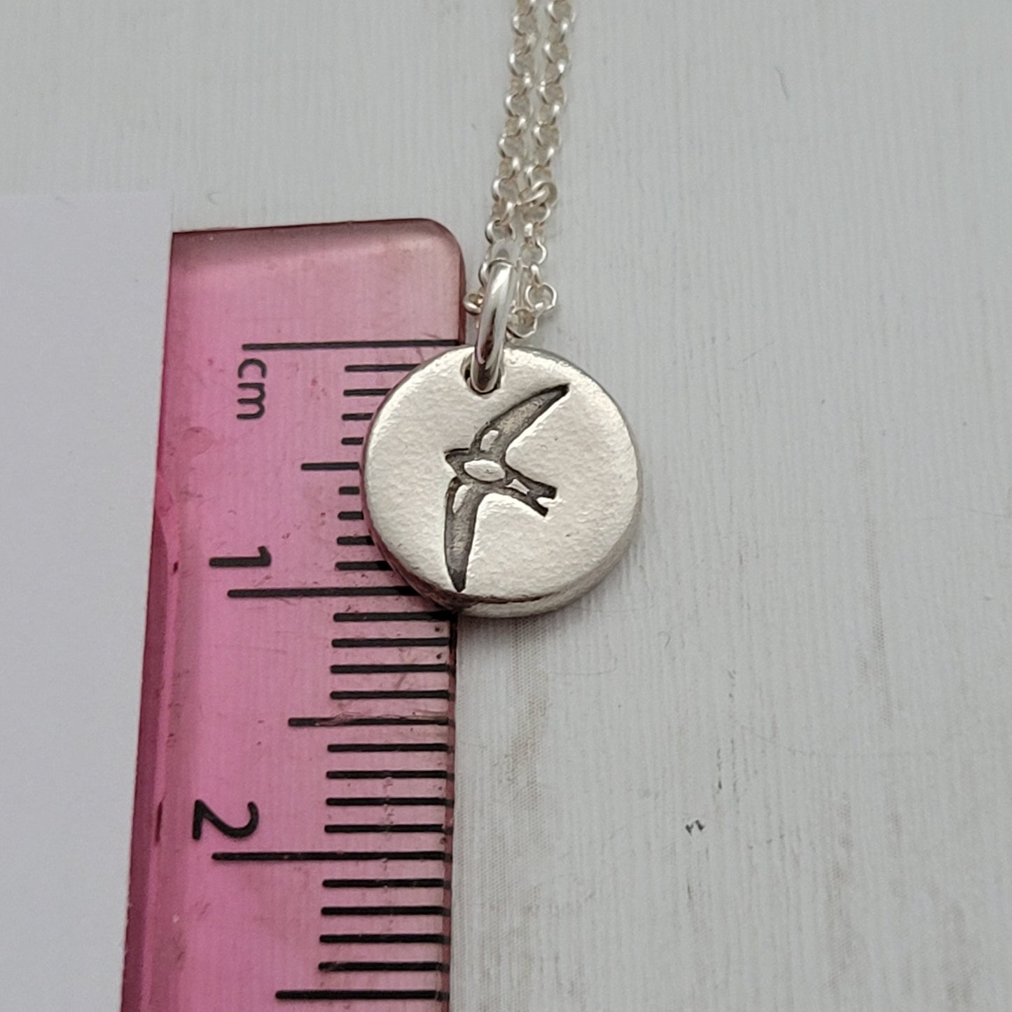 Round silver bird design pendant necklace next to ruler