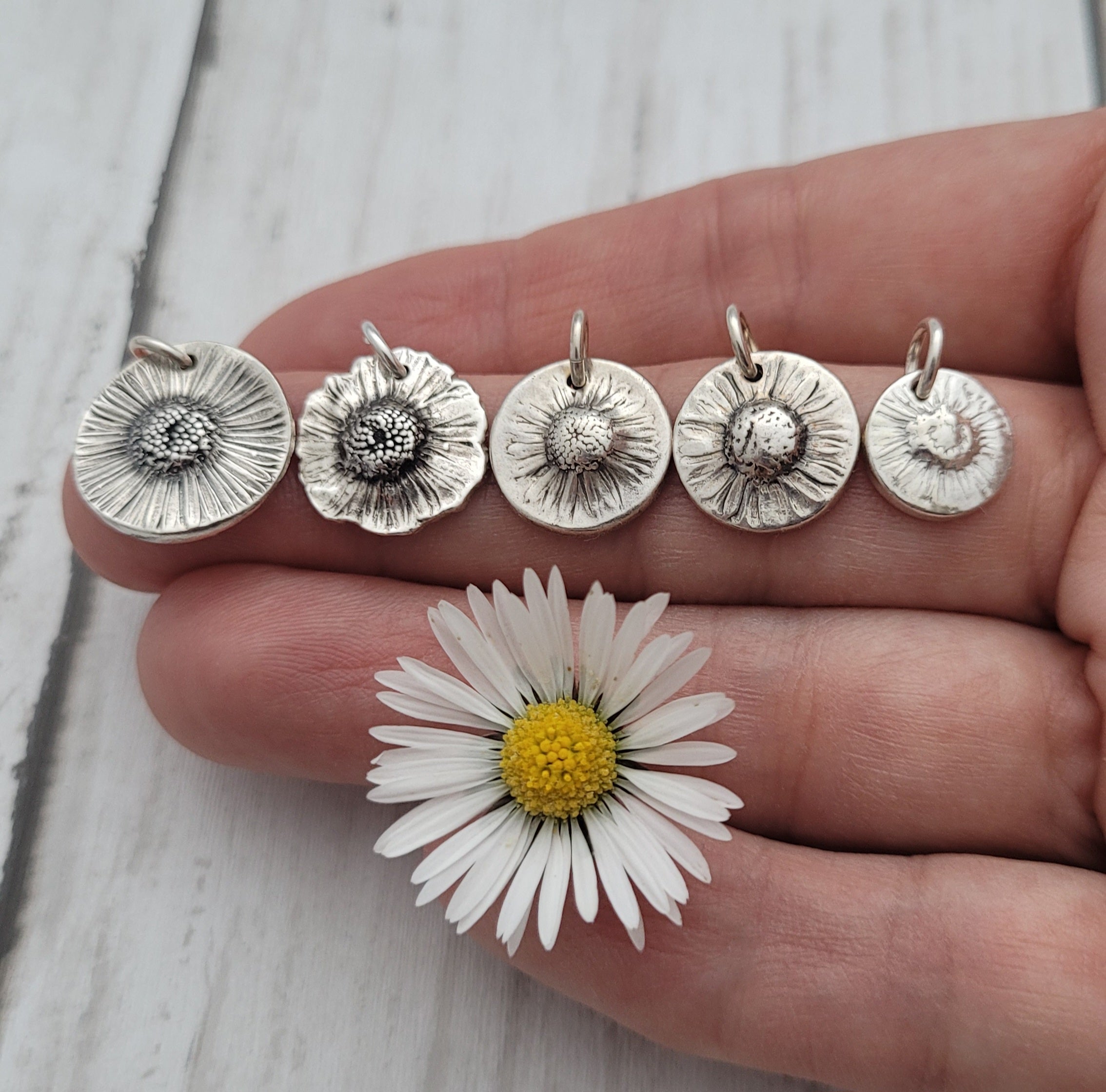 Five silver flower pendants on a hand next to a daisy flower.