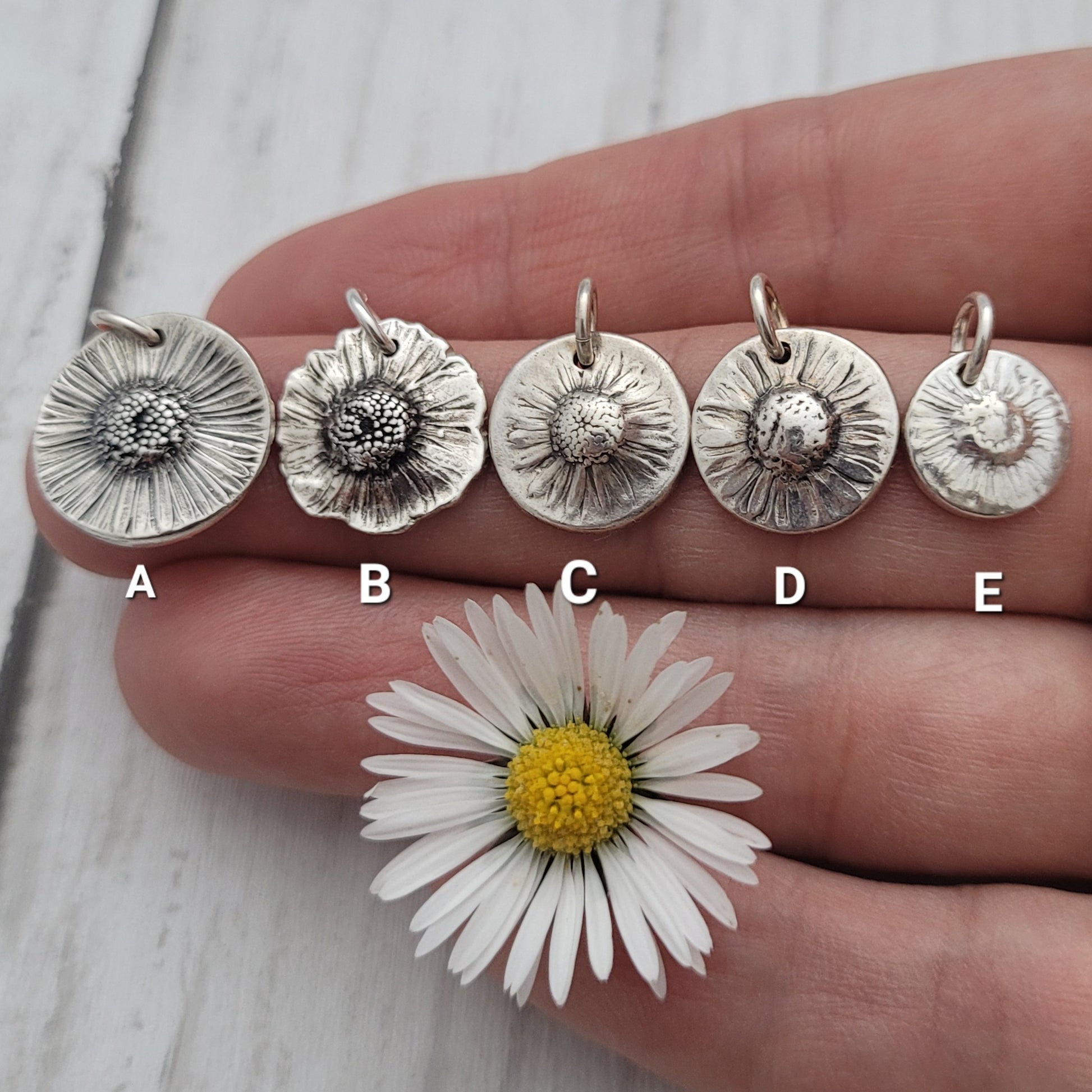 Five silver flower pendants on a hand with a daisy flower for size comparison.