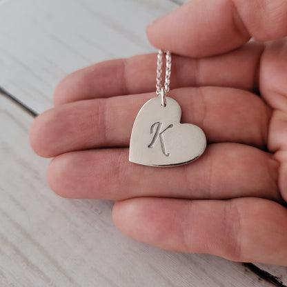 Silver heart-shaped pendant with 'K' held in a hand on a light wooden surface
