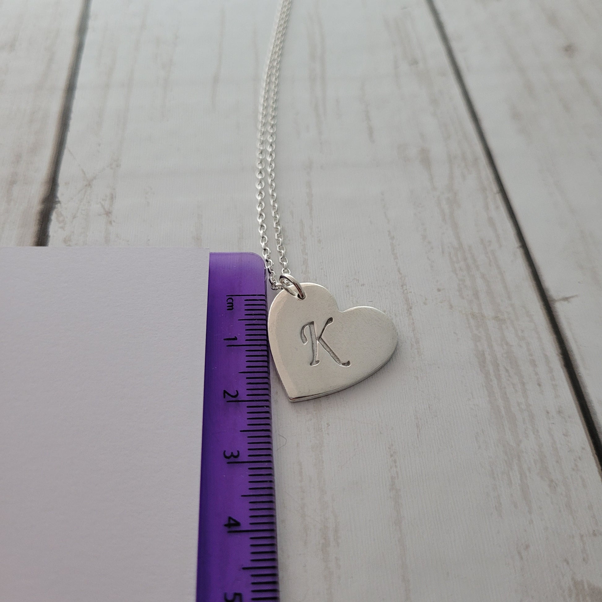 Silver heart-shaped pendant with engraved design on a wooden surface with a ruler for scale.