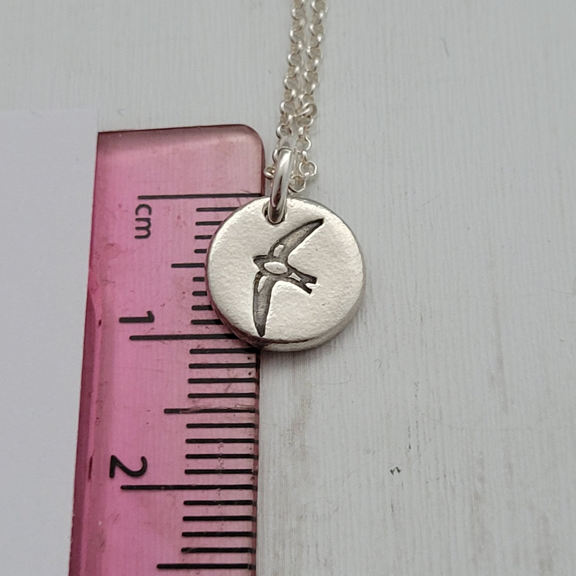 Round silver bird design pendant necklace next to ruler