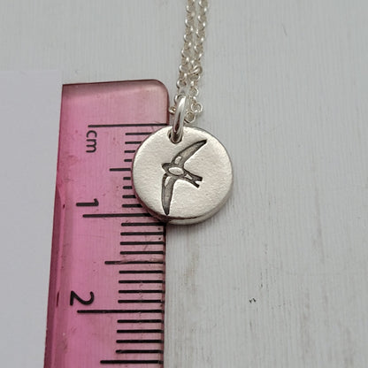 Round silver bird design pendant necklace next to ruler