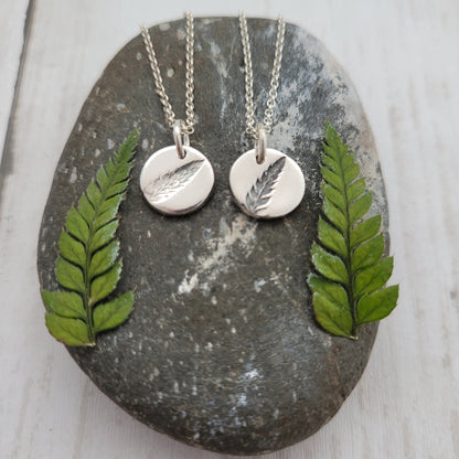 Two silver necklaces with leaf pendants on a stone surface with green leaves.