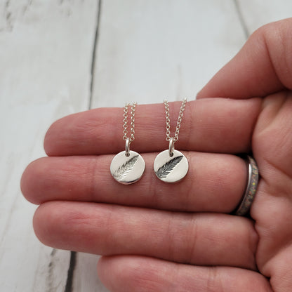 Two silver necklaces with fern pendants on a person's hand against a white background
