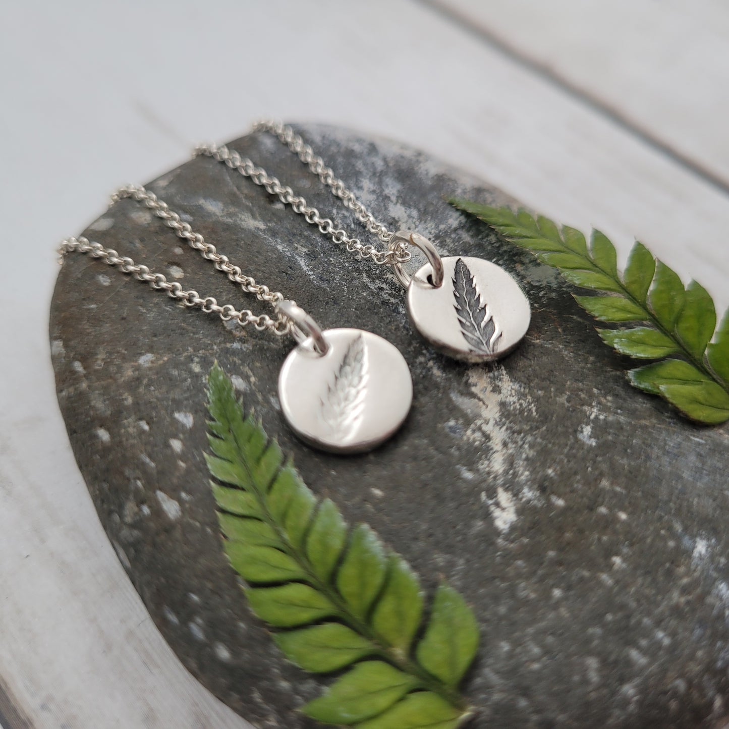 Silver necklaces with leaf pendants on a stone surface with green leaves.