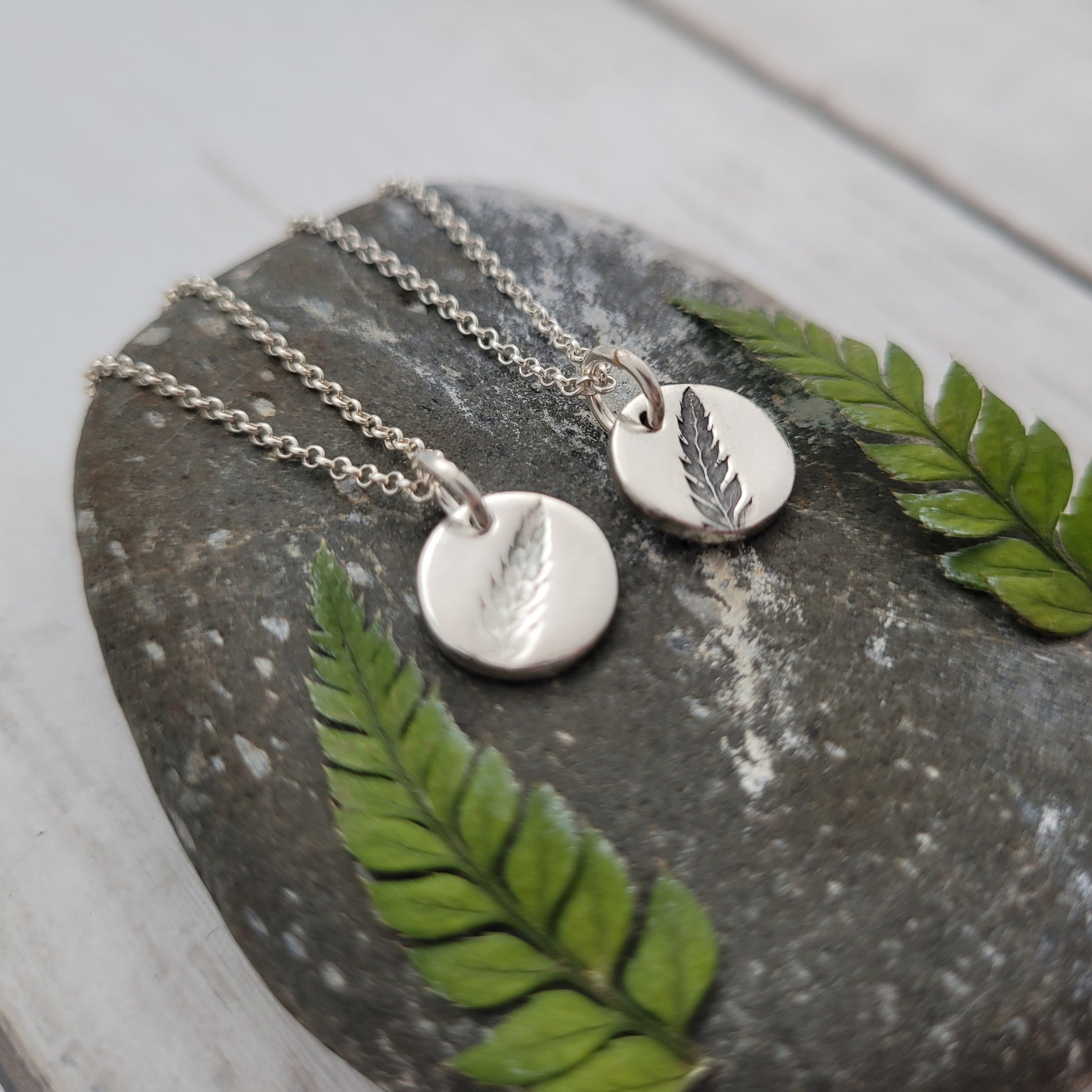 Silver necklaces with leaf pendants on a stone surface with green leaves.