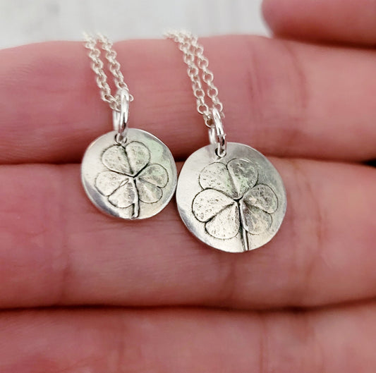 Two silver shamrock pendants on chains held between fingers.