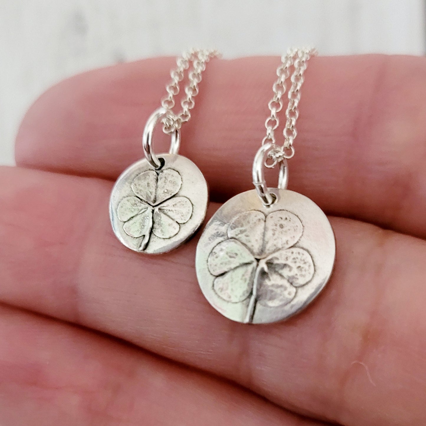 Two silver four-leaf clover pendants on chains held between fingers against a neutral background