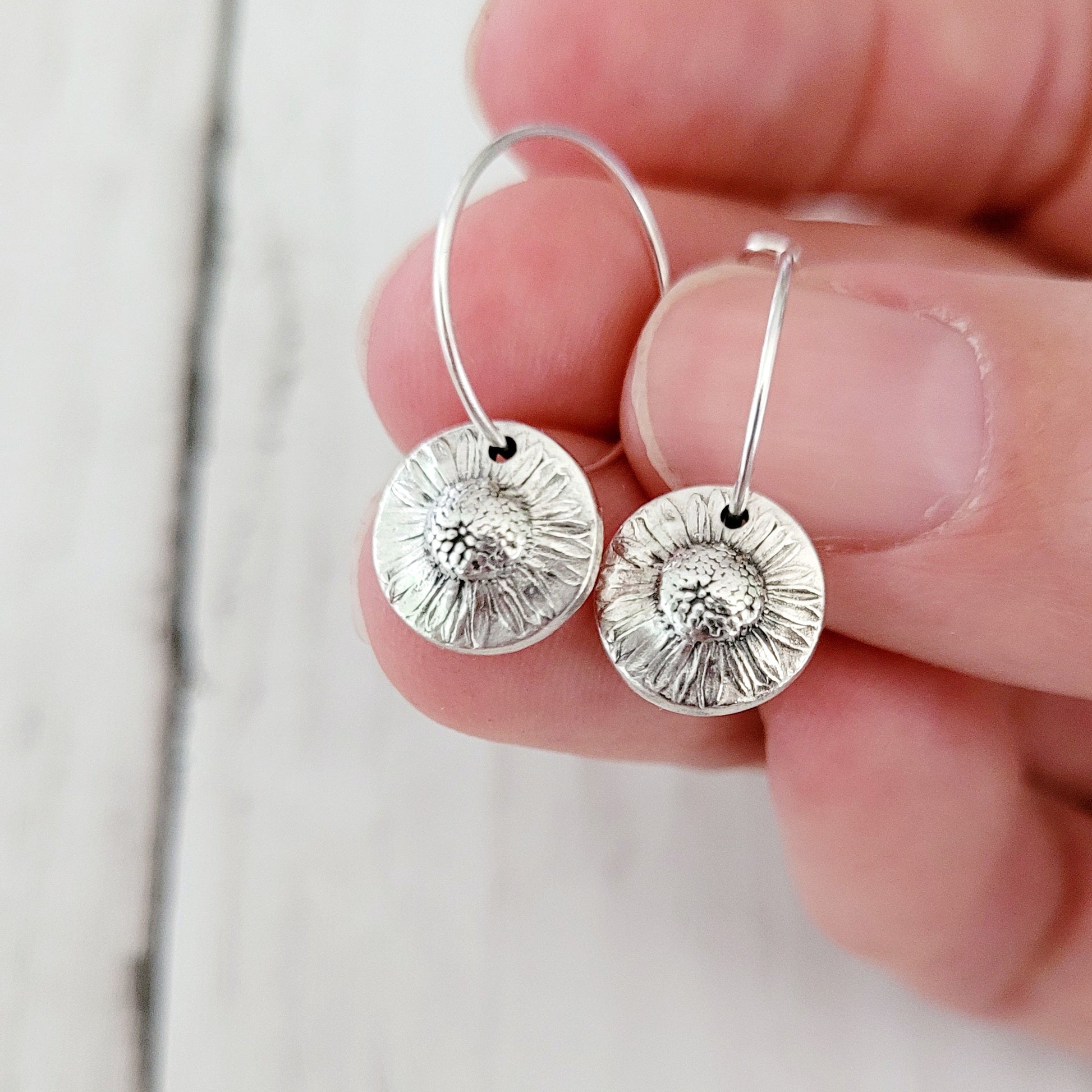 Silver daisy hoop earrings held between fingers against a light background
