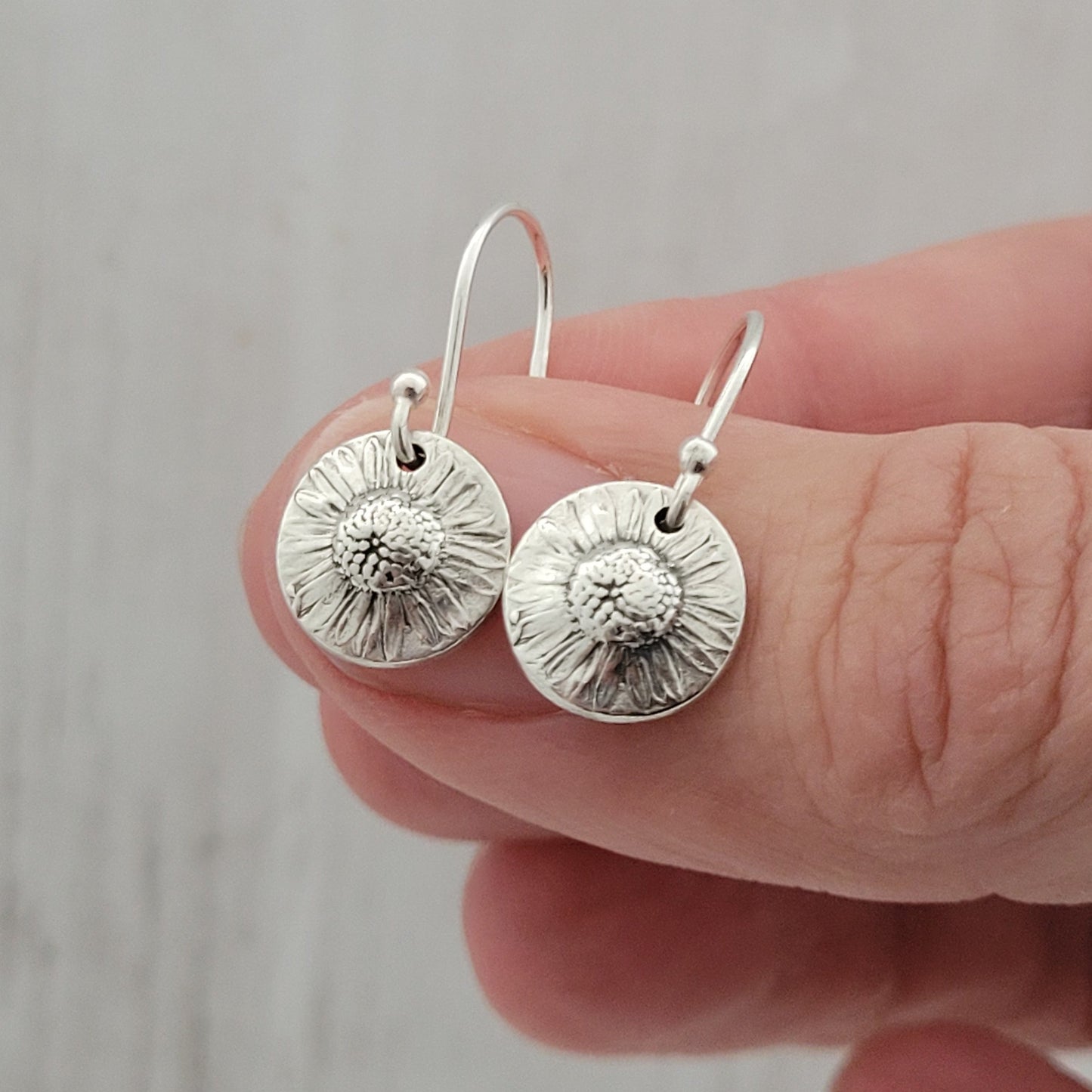Silver daisy earrings held between fingers against a neutral background