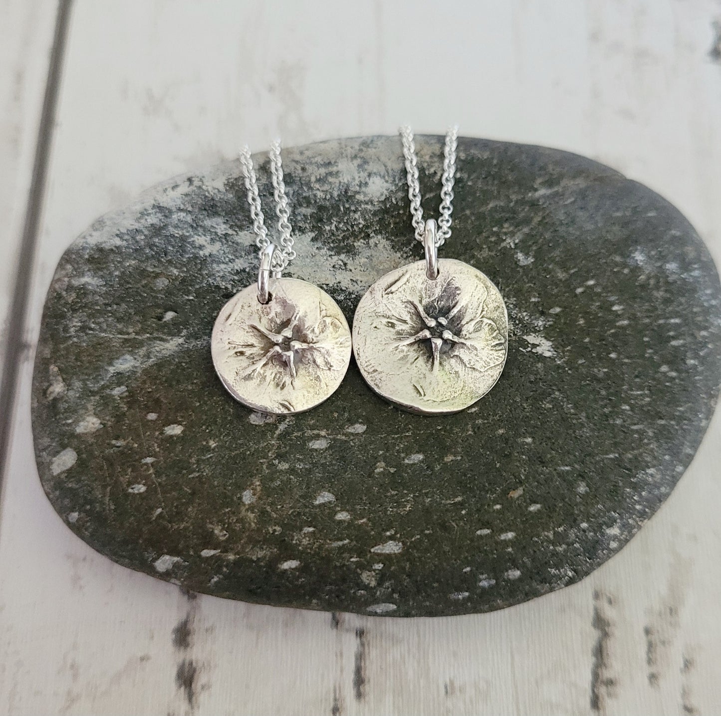 Two silver circular  green alkanet flower pendants on chains resting on a dark stone surface.