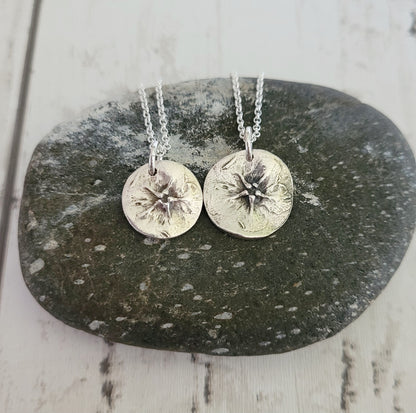Two silver circular  green alkanet flower pendants on chains resting on a dark stone surface.