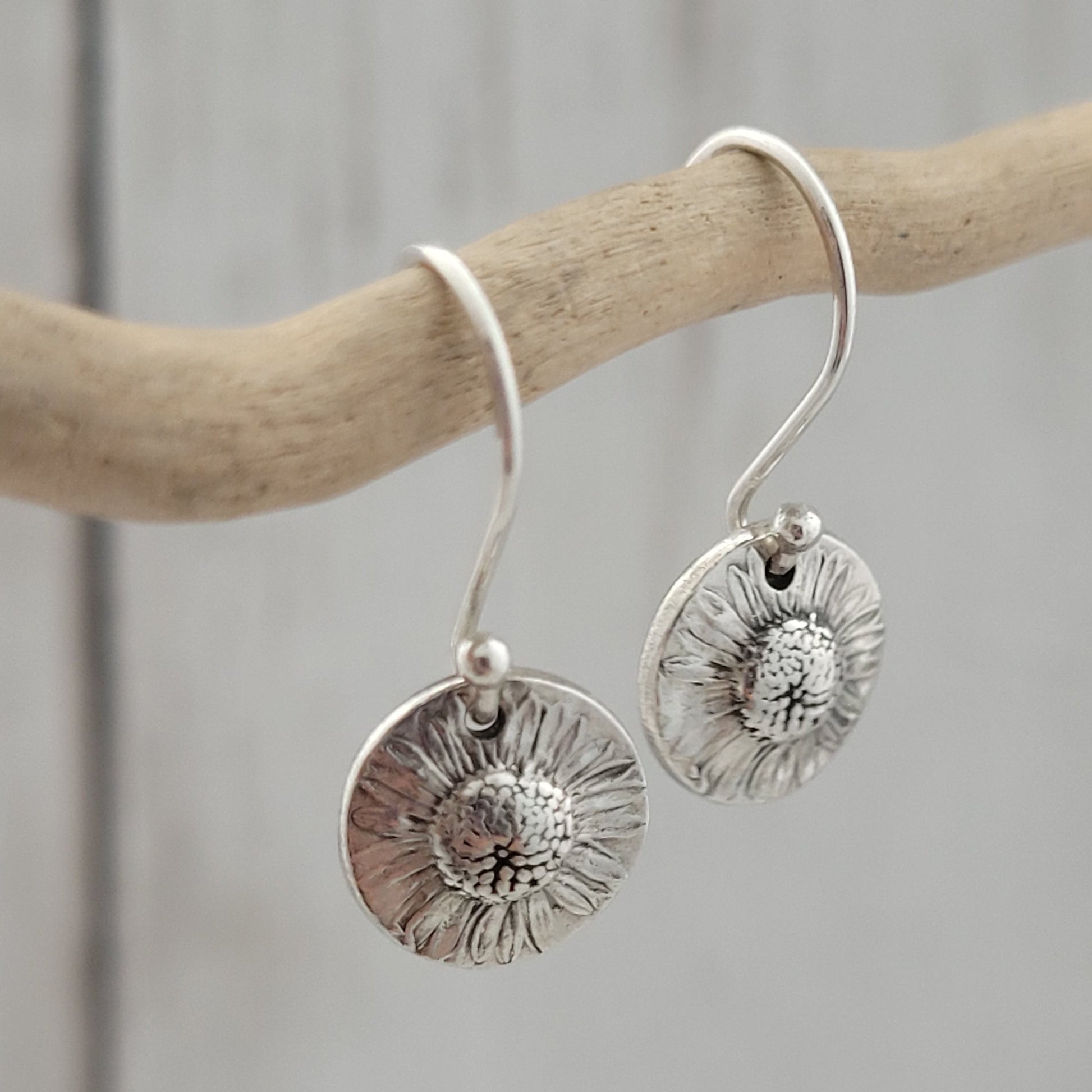 Silver floral earrings hanging from a branch on a neutral background