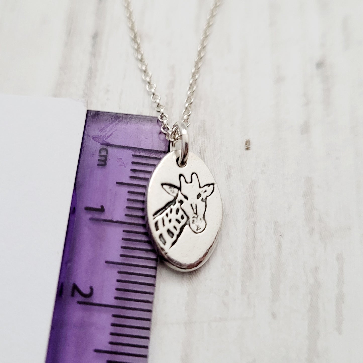Silver necklace with a giraffe pendant next to a ruler on a white background