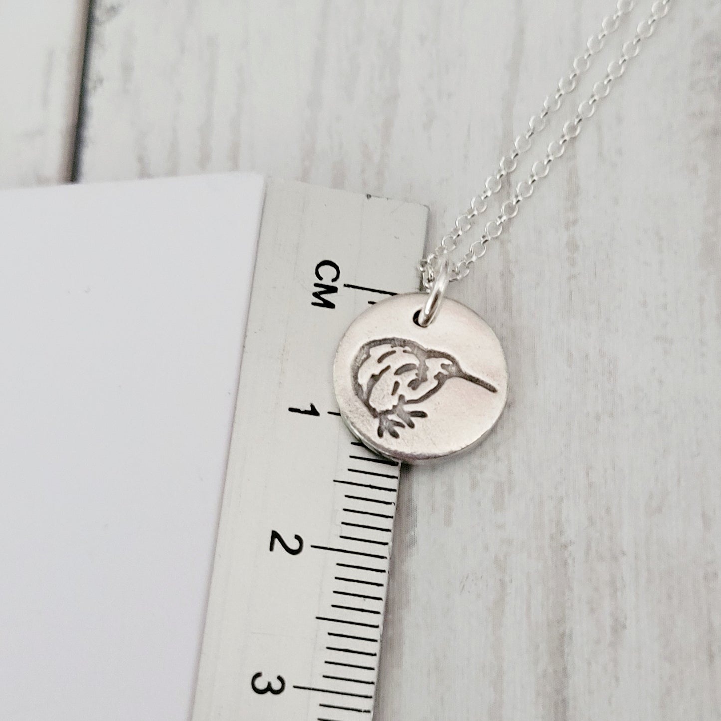Silver necklace with a kiwi bird pendant on a ruler for scale